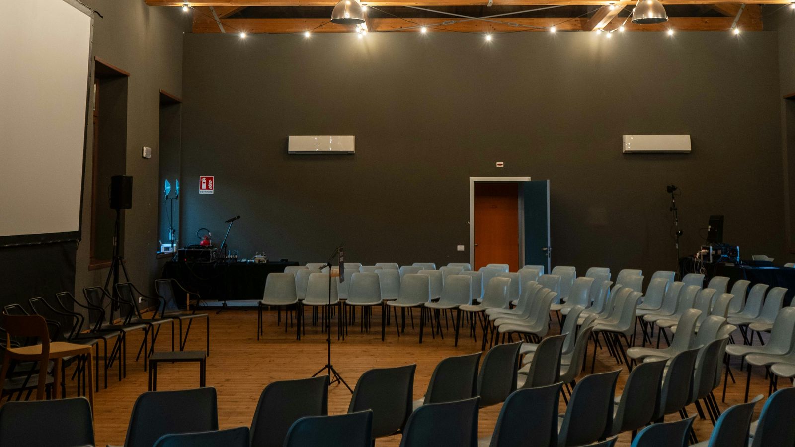 An empty meeting room with rows of gray chairs curved around an open wooden floor, under wooden ceiling beams and string lights.