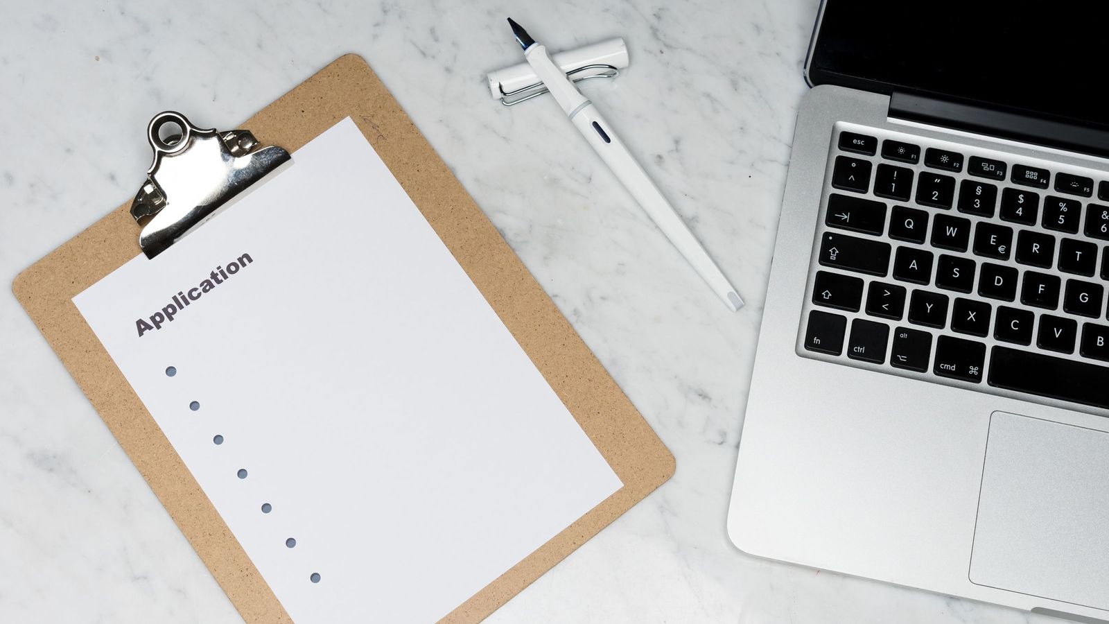 A blank application form on a clipboard beside a white pen and an open laptop on a marble desk.