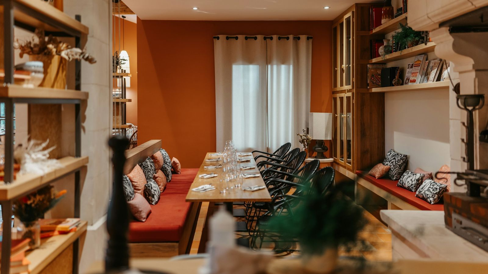 A long wooden table set for a shared meal, with empty chairs and bench seating in a warm, orange-walled room.